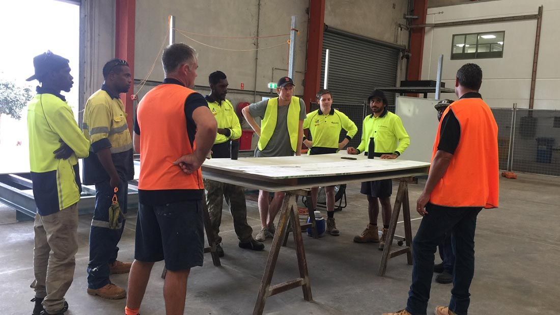 Group of workers in safety vests gather around a table inside a warehouse, discussing a project.