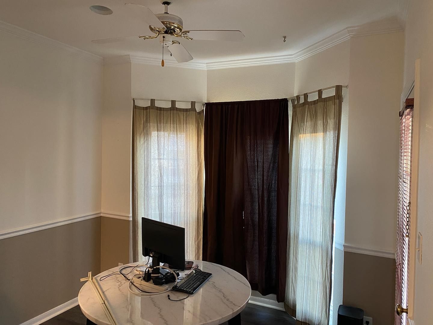 A round desk with a computer and curtains in a room with a ceiling fan.