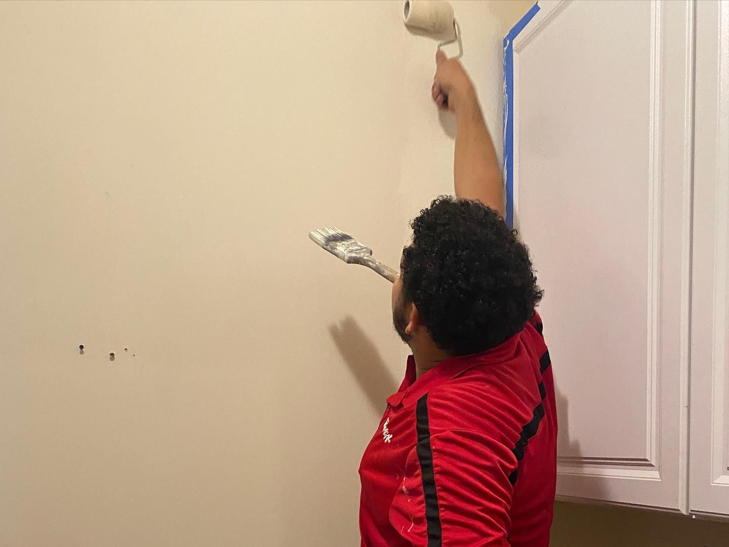Person painting a wall with a roller. They are wearing a red shirt and holding a putty knife.