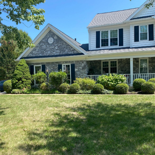 Two-story house with stone facade, dark shutters, white trim, and a porch on a sunny day.