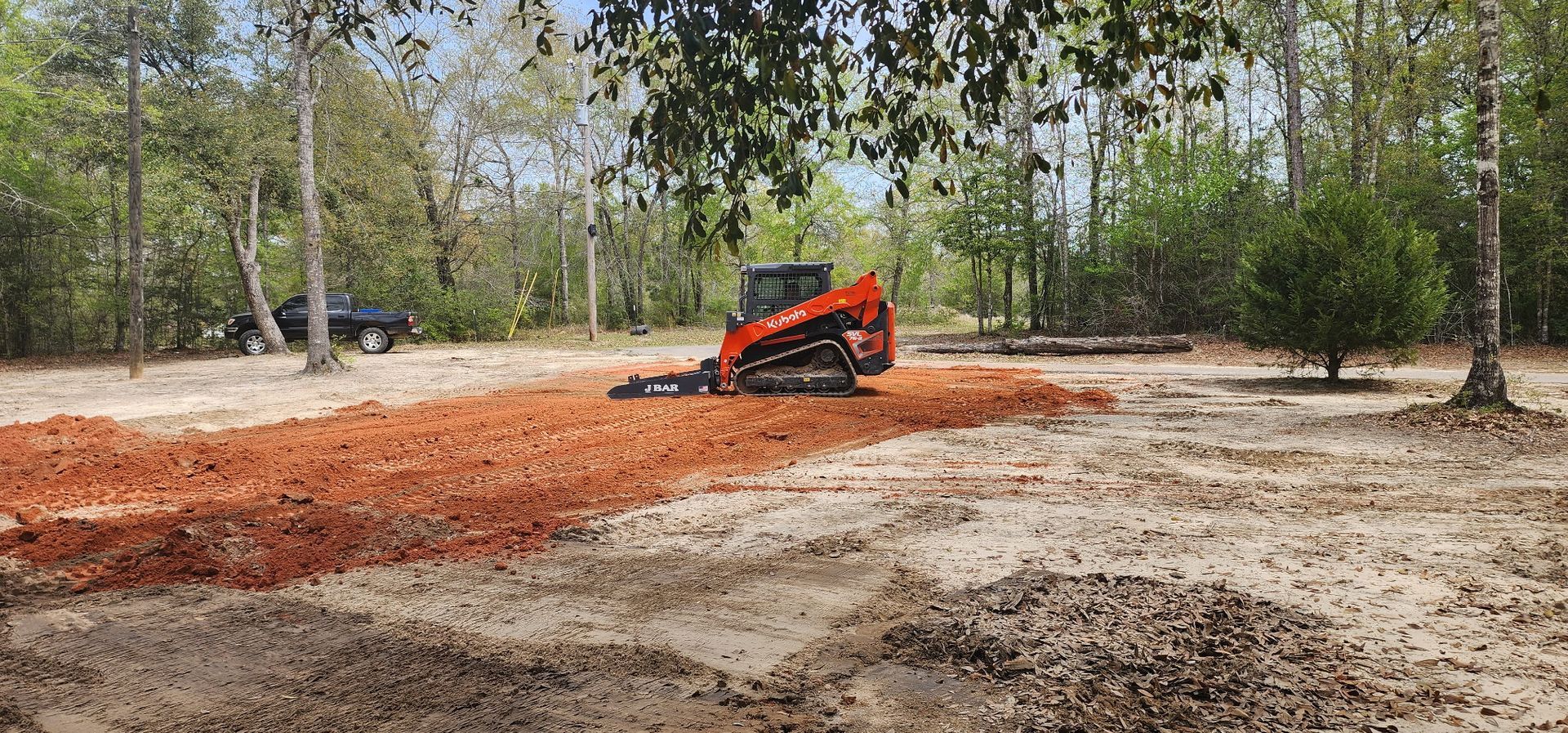 An orange skid steer tractor is grading red soil in a wooded area. A car is visible in the background.