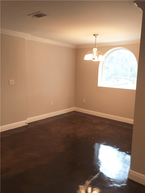 Empty room with brown floor, tan walls, white trim and chandelier. Window on right.