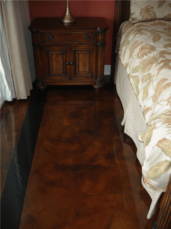 Brown stained concrete floor in a bedroom, with a wooden nightstand and part of a bed.