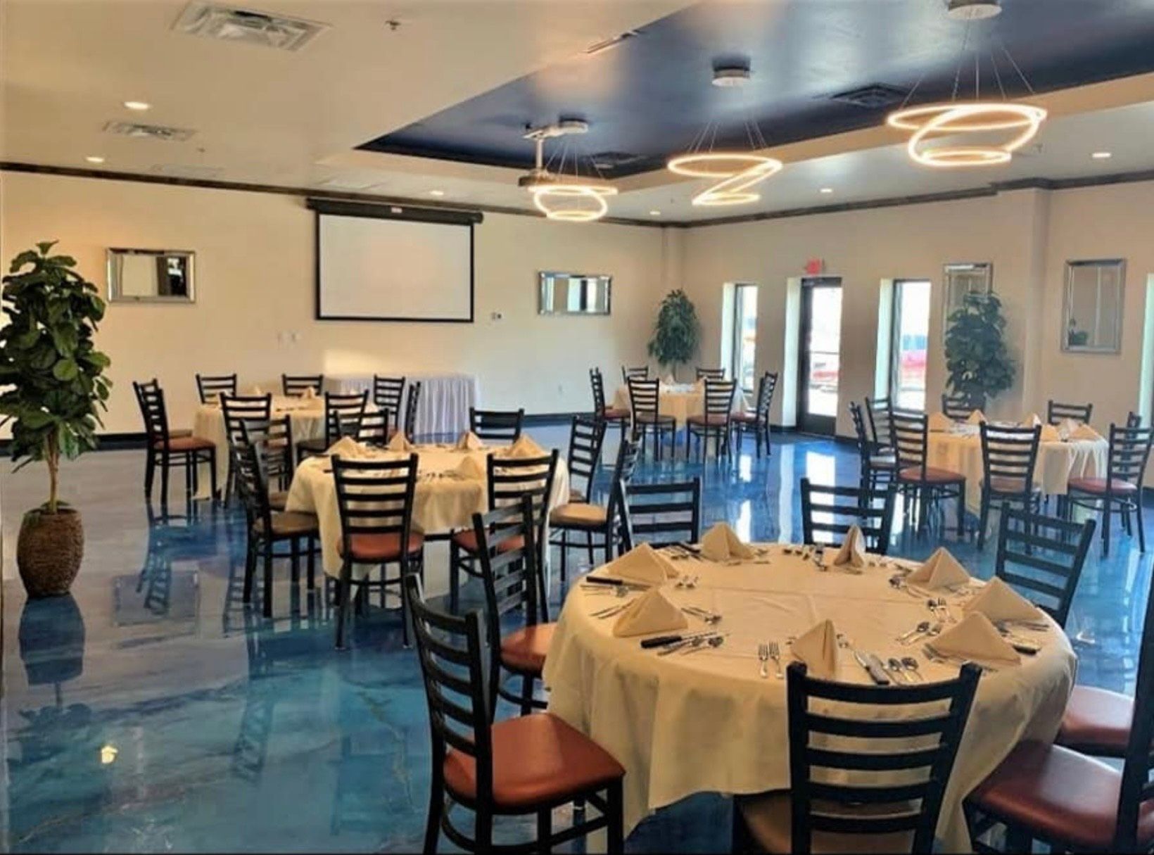 Banquet hall with round tables set for dining, blue floor, projection screen, and decorative lighting.