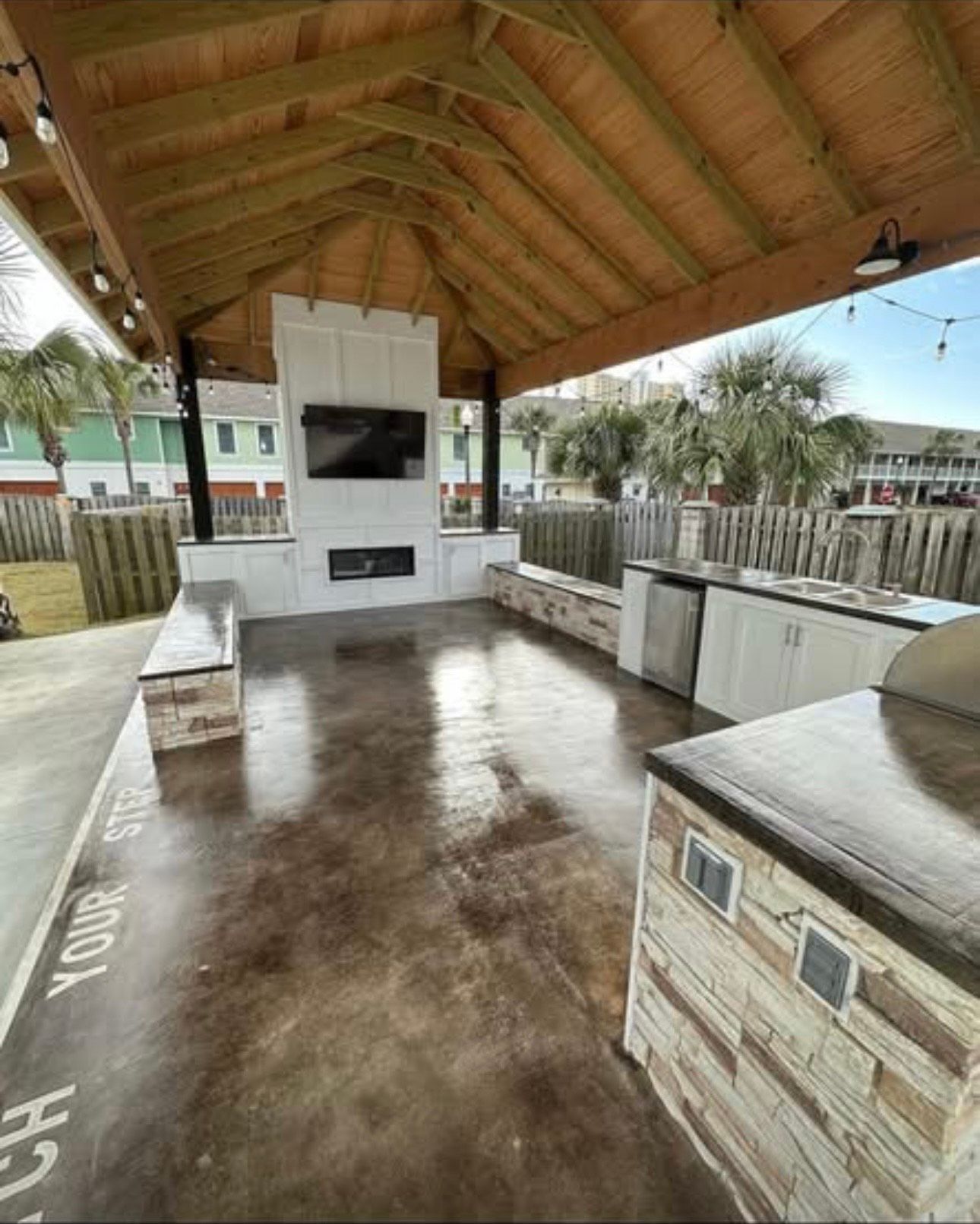 Outdoor kitchen area with a TV, fireplace, countertops, and a wooden roof.