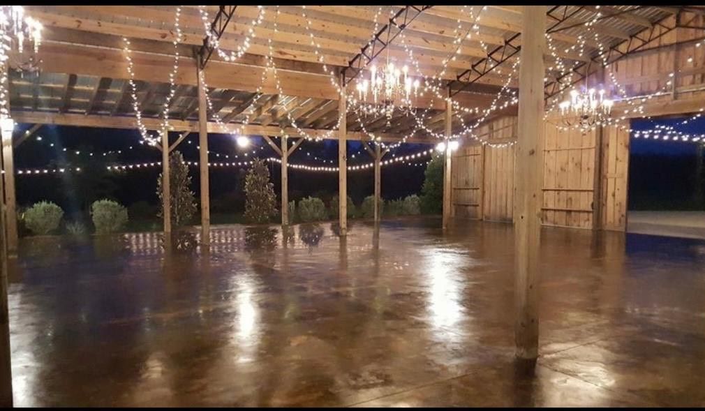 Inside of barn with string lights, polished floor, and outside view of bushes and trees at dusk.