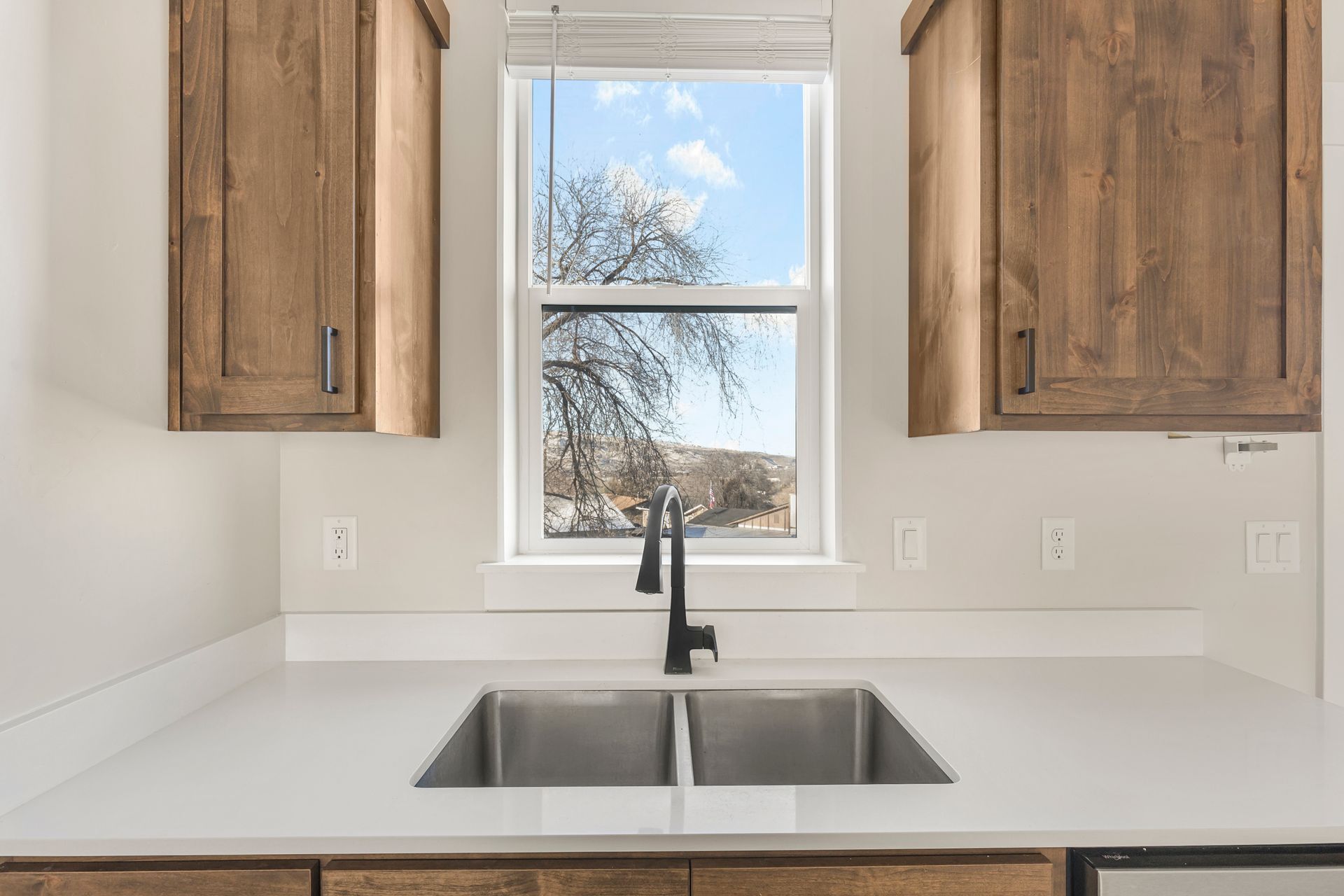 Kitchen with a double sink, black faucet, white countertop, and wooden cabinets; window shows a tree and sky.