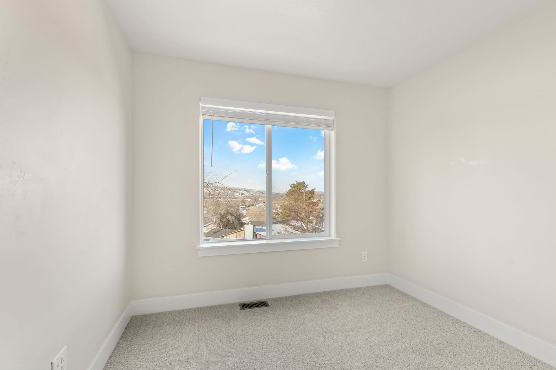 Empty room with a window, white walls, light carpet, and a view of a town under a blue sky.