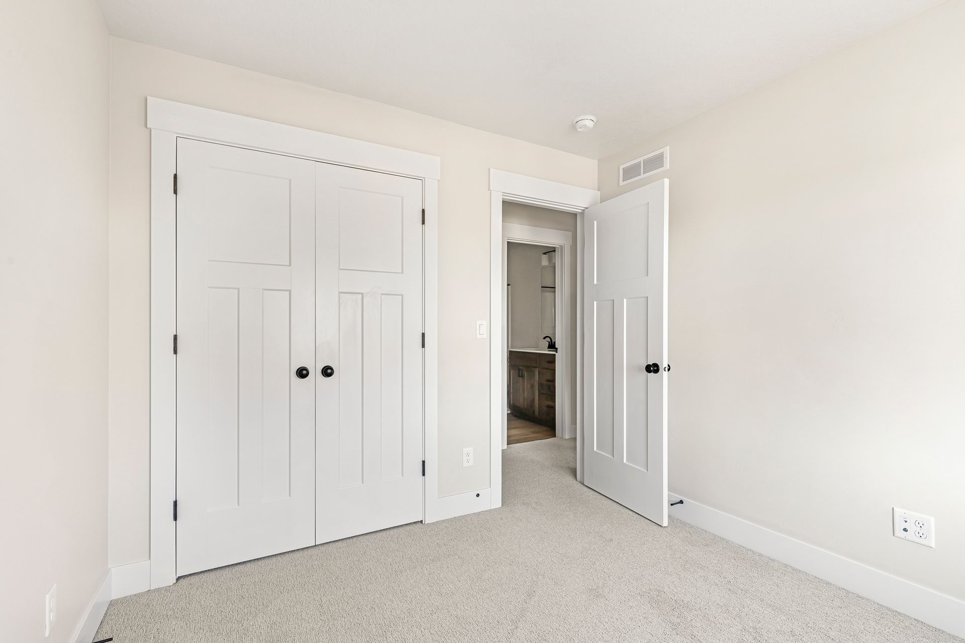 Empty bedroom with white doors, closet, and a doorway to a bathroom. Beige walls and carpet.