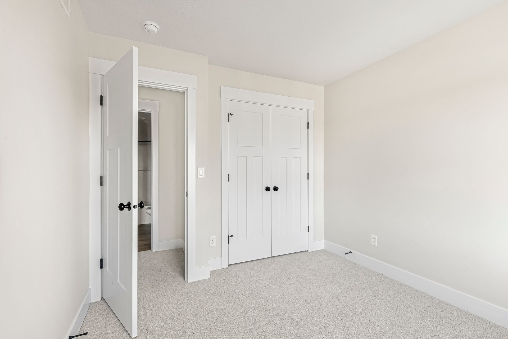 Empty bedroom with white walls, carpet, and closed white closet doors. One door is ajar.