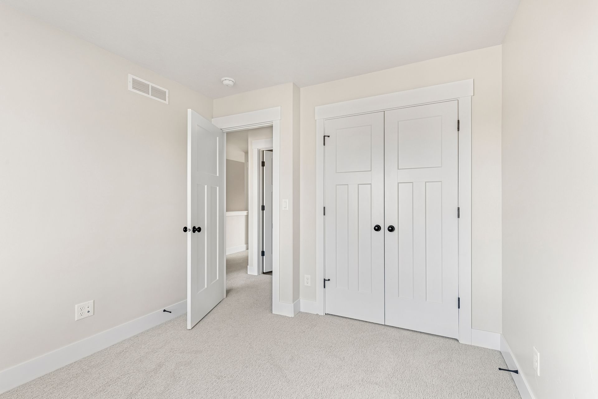 Empty bedroom with white walls, carpet, and closed closet doors. An open doorway leads to a hallway.