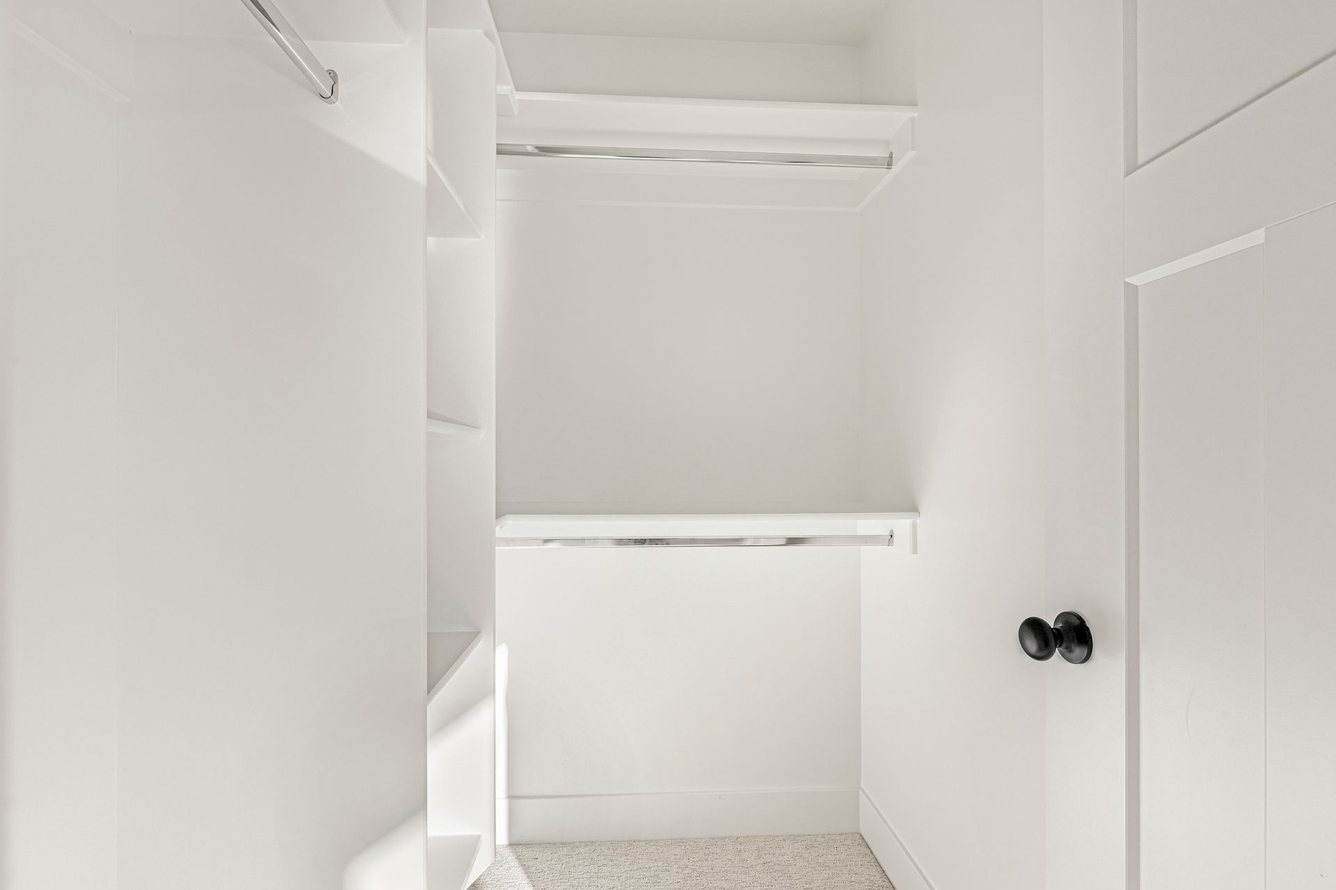 Empty white closet with a rod and shelf. A closed door with a black handle is on the right.
