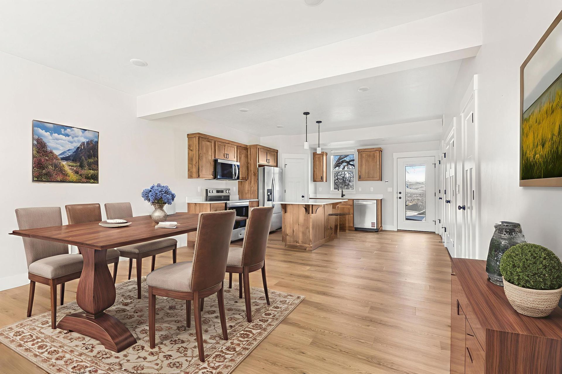 Dining area with wooden table, chairs, kitchen, and art on white walls.