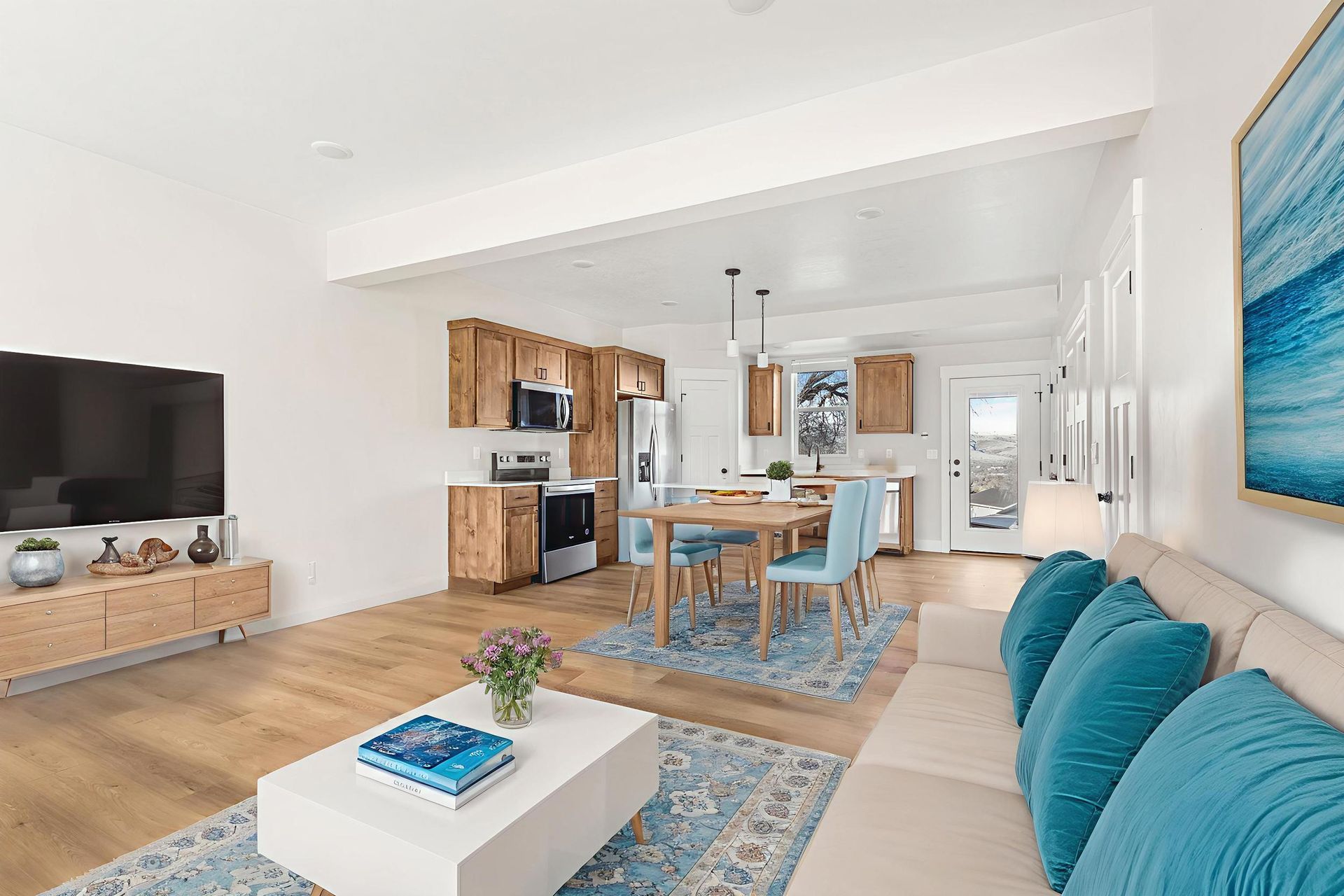 Open-concept living space with kitchen, dining area, and living room; light wood floors, blue accents, and white walls.