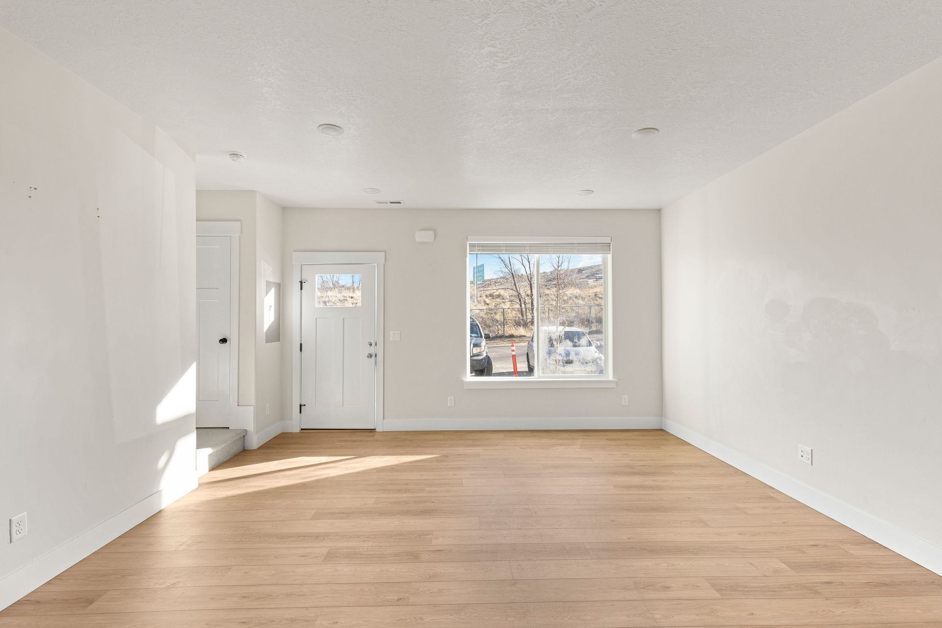 Empty living room with light wood floors, white walls, door, and window.