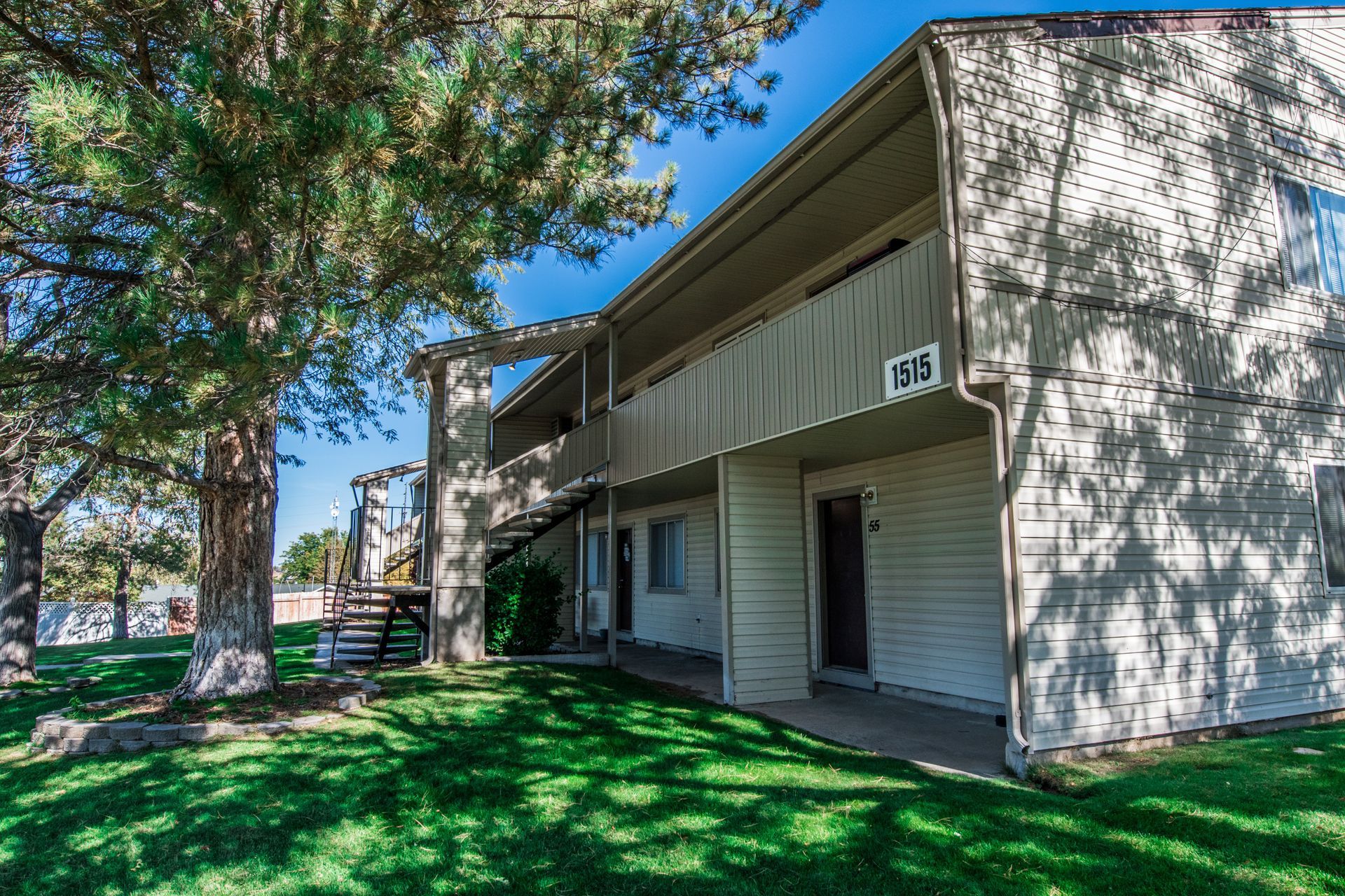 Two-story beige apartment building with balconies, green lawn, and a tree on a sunny day.