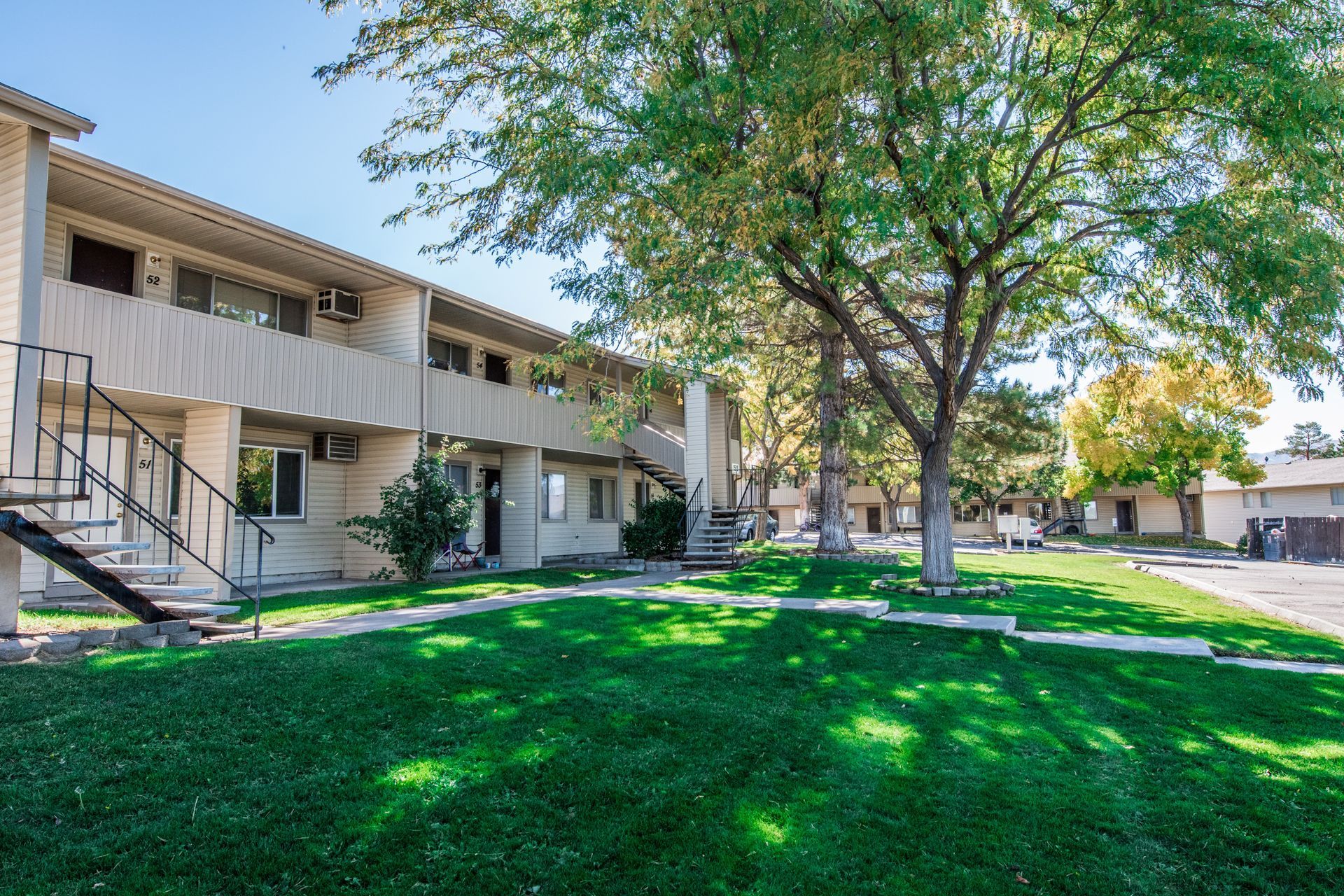Two-story beige apartment building with green lawn and trees on a sunny day.