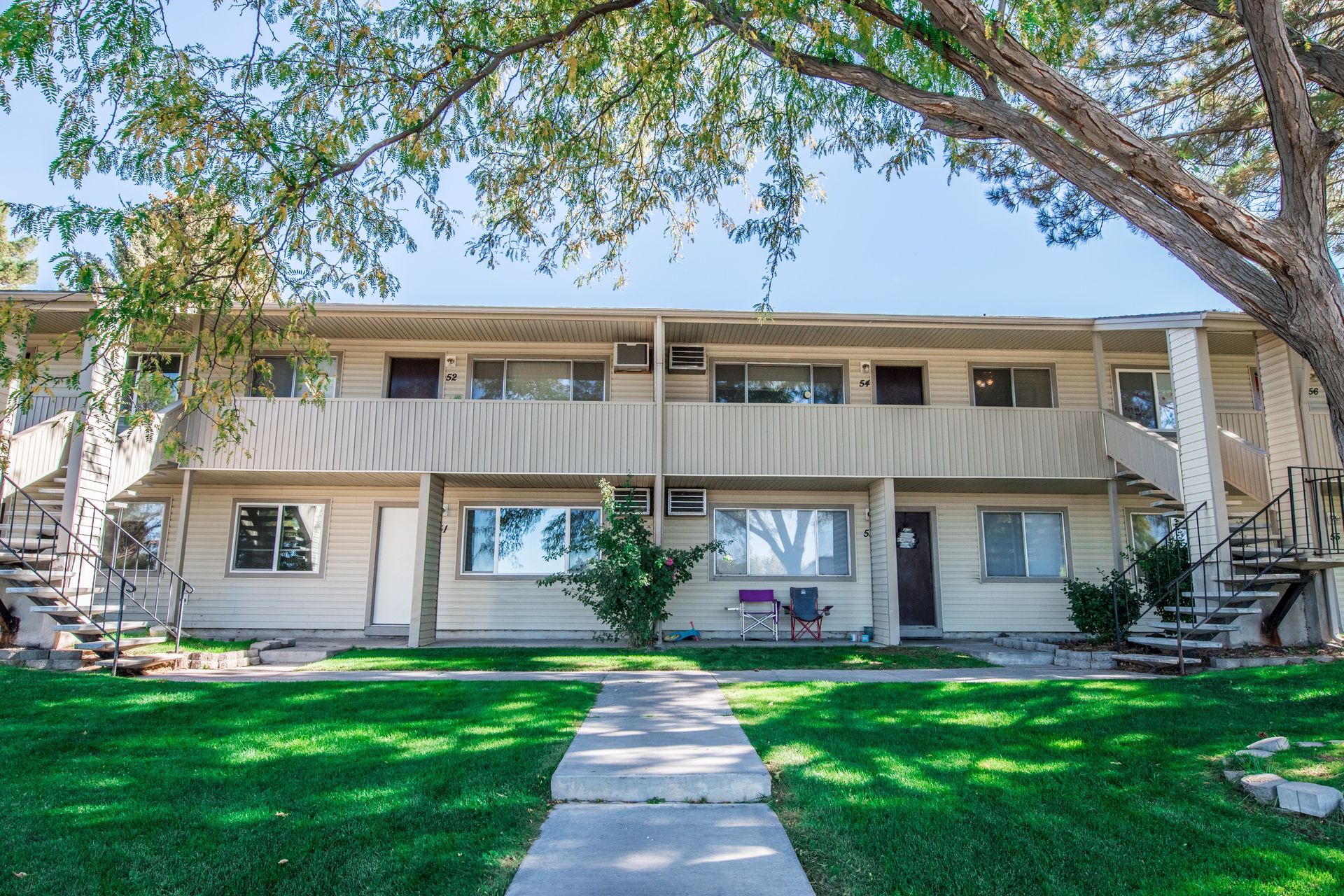 Two-story apartment building with green lawn and walkway under a tree on a sunny day.