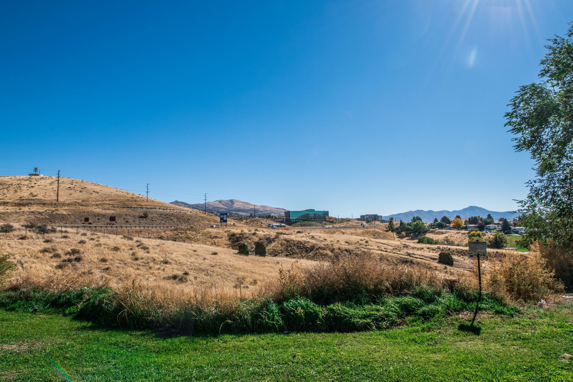 Grassy field with dry vegetation and a blue sky; distant mountains and buildings.