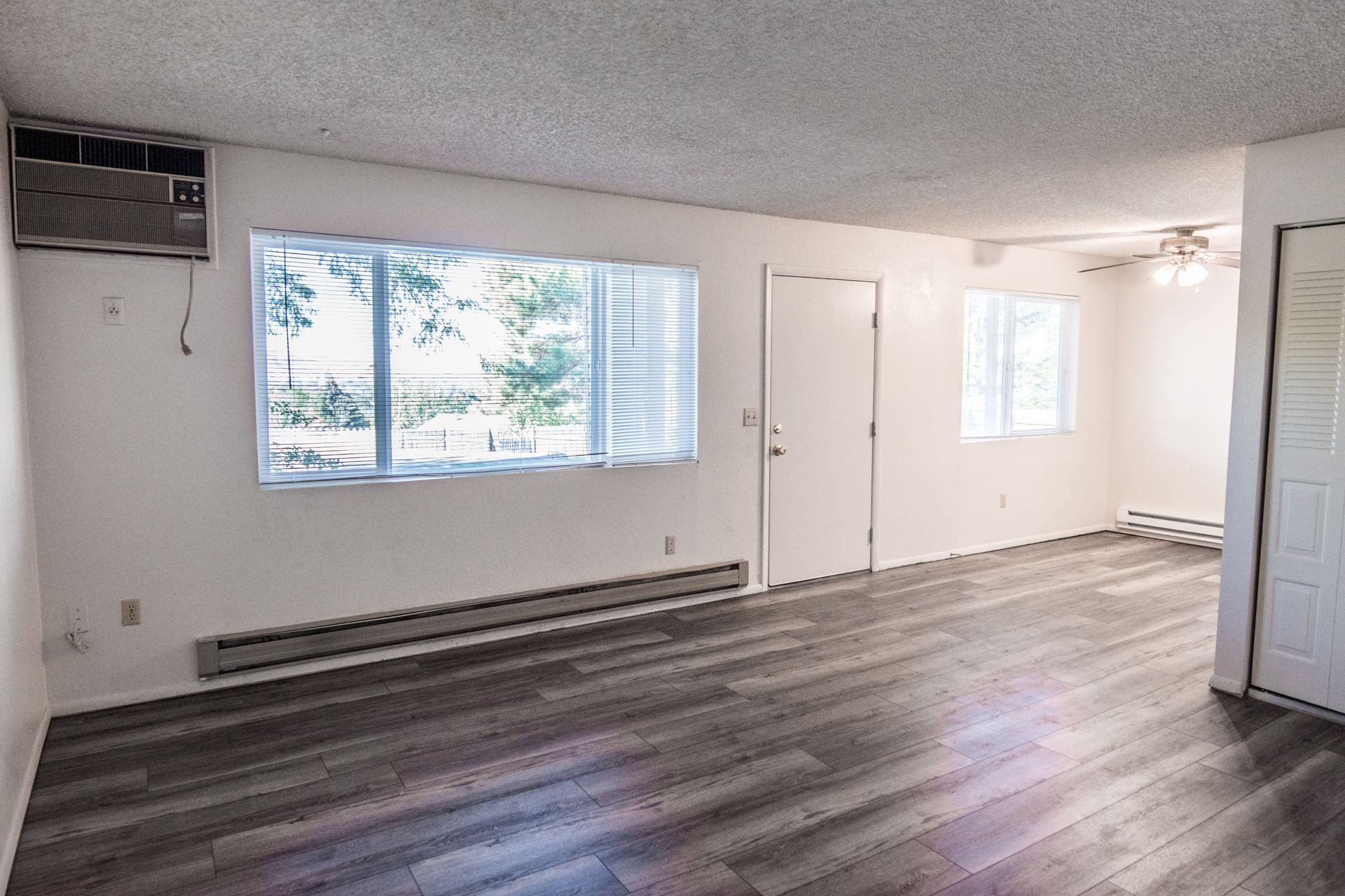 Empty, bright apartment interior with large window, gray flooring, white walls, and an air conditioner.