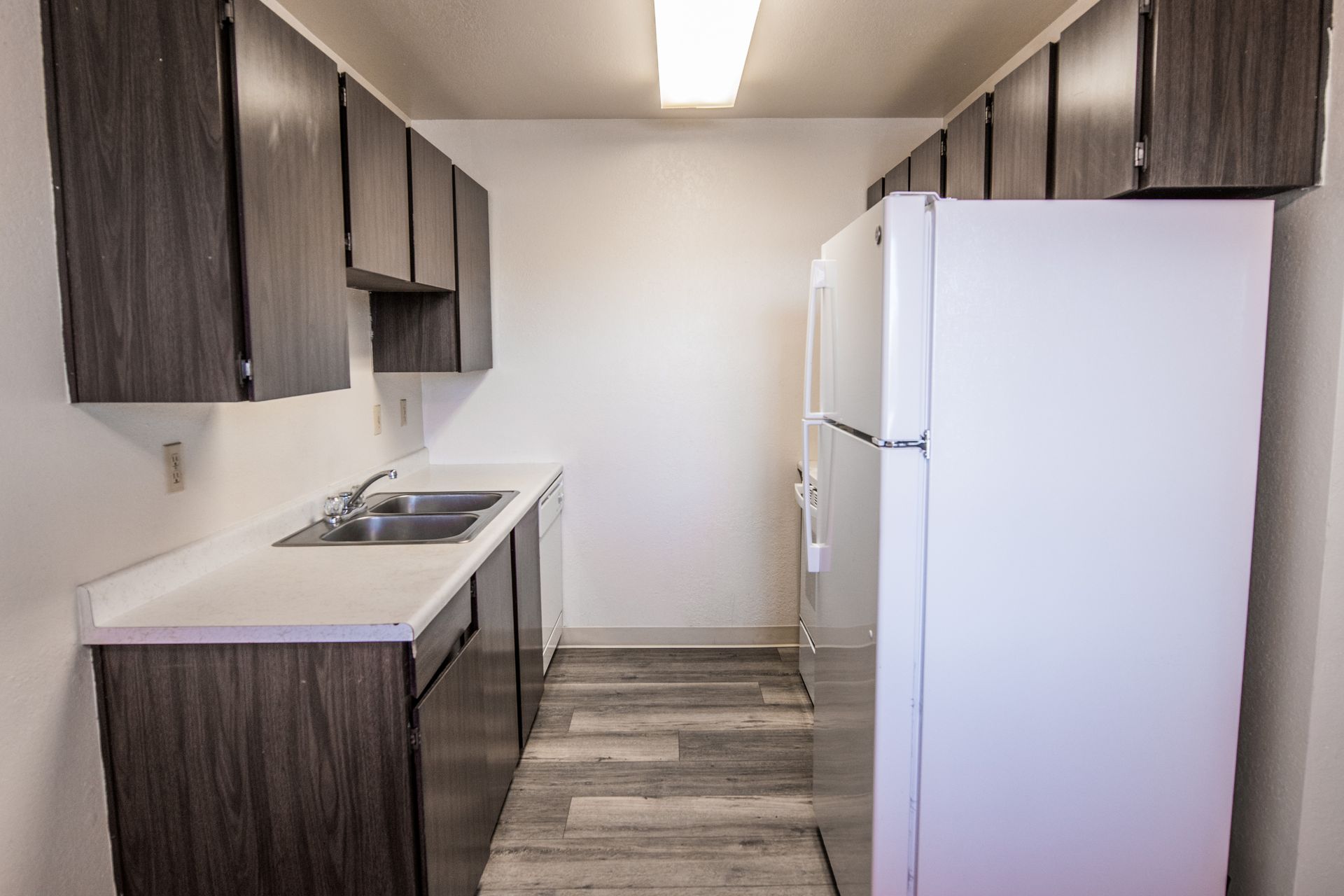 A narrow kitchen with dark cabinets, white countertops, and a white refrigerator; gray flooring.