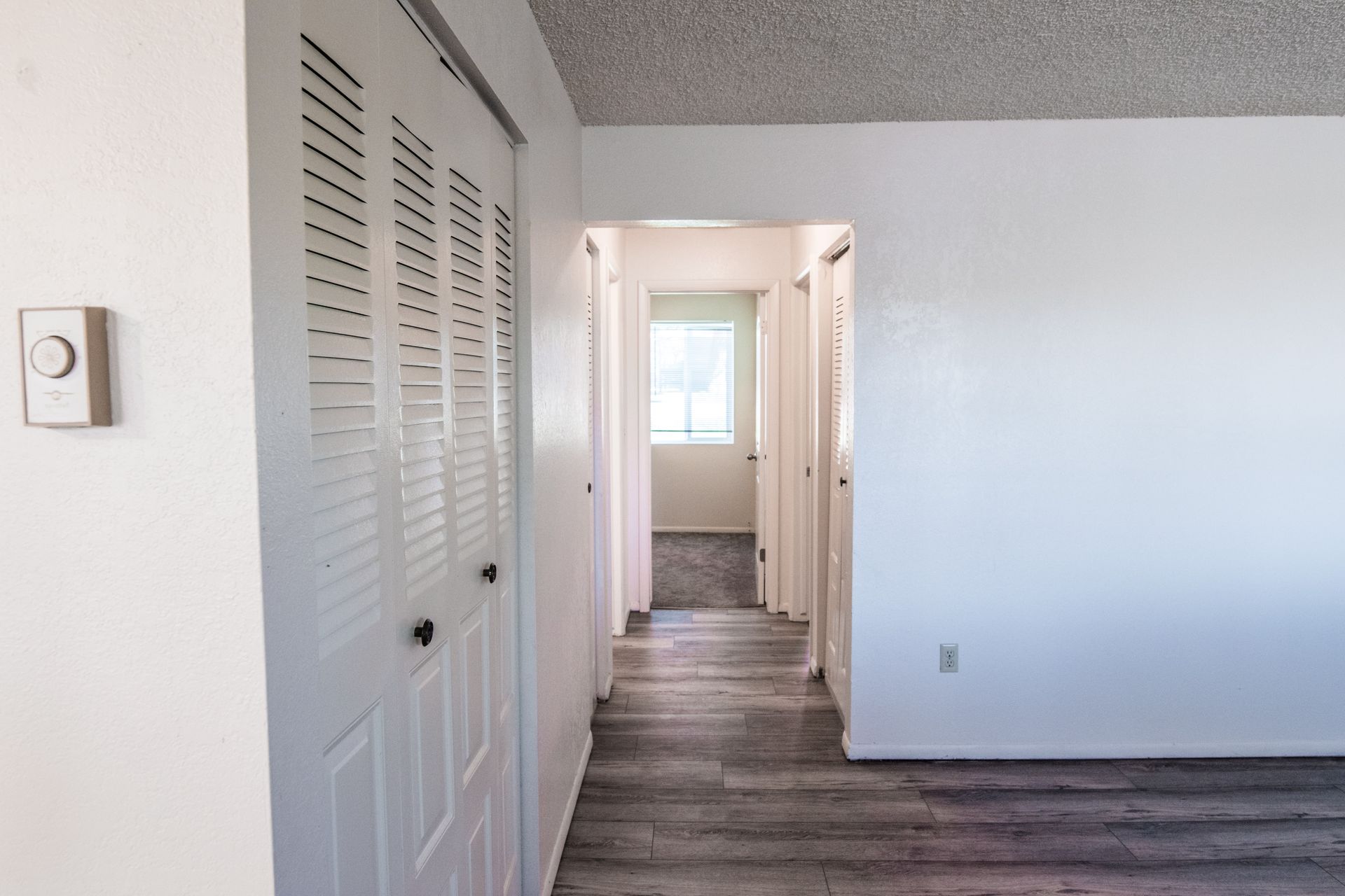 Hallway with closet doors and a doorway to a room with a window; white walls, gray flooring.