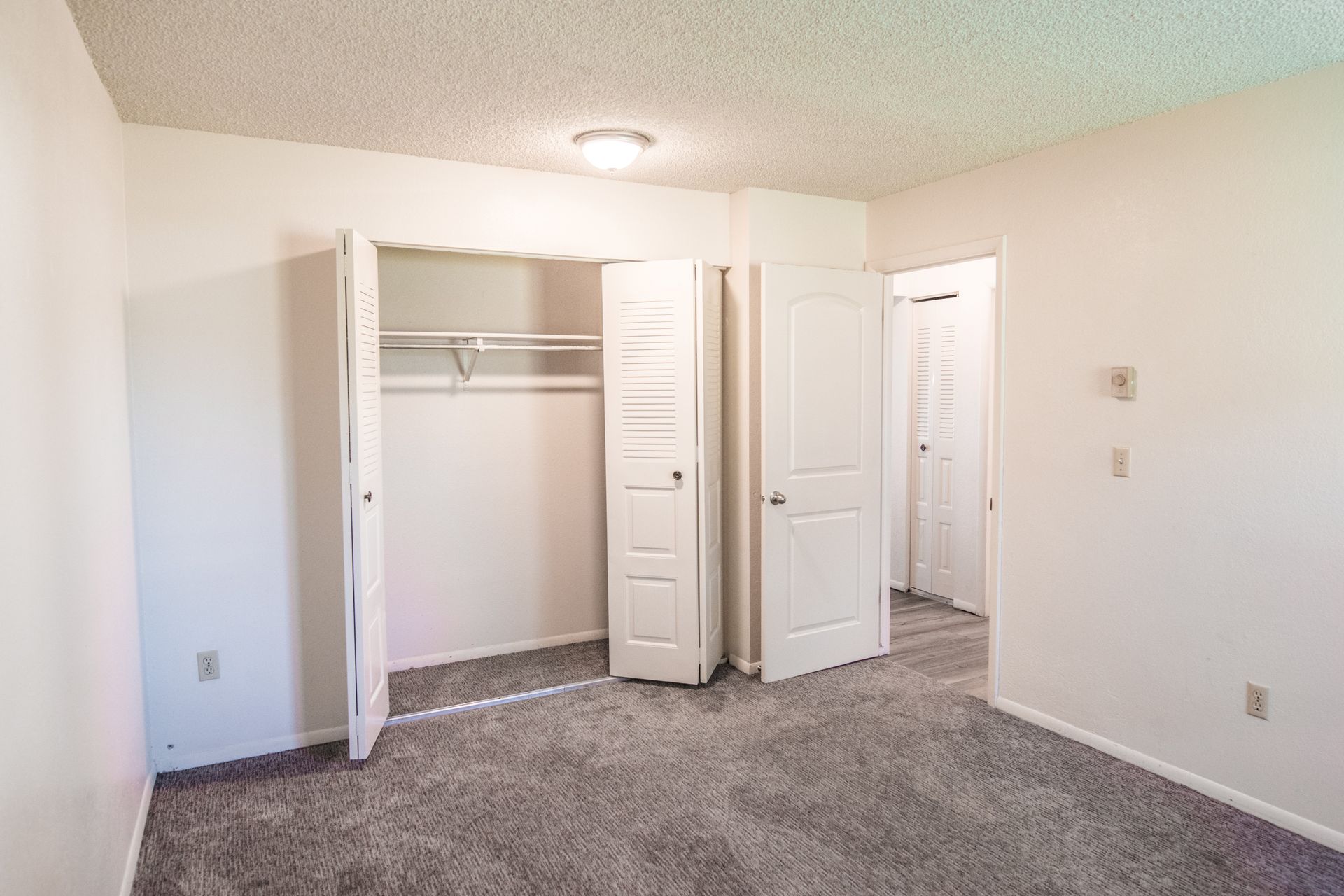 Empty bedroom with a closet and a doorway, gray carpet, white walls, and a popcorn ceiling.