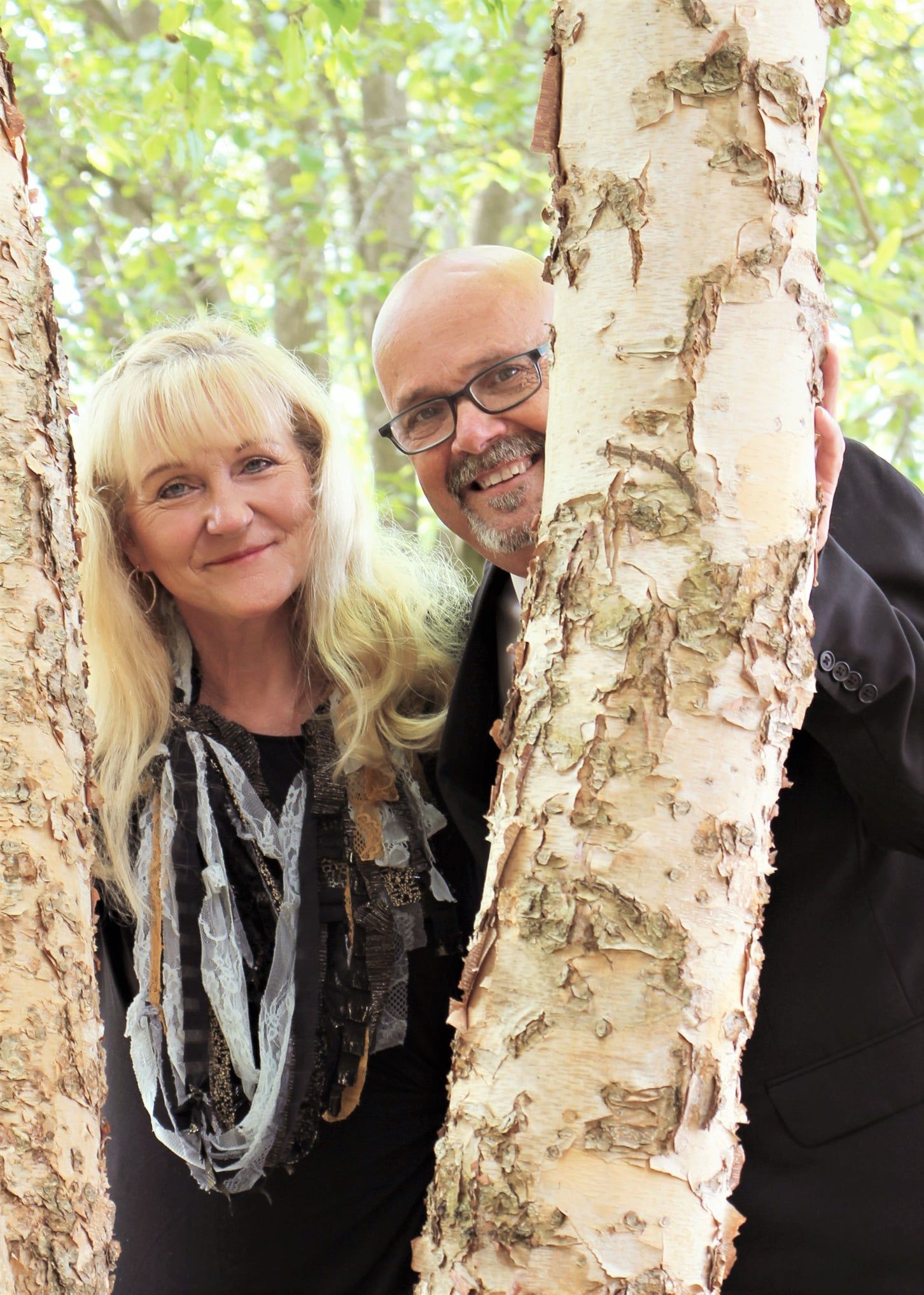 Woman and man smiling, peeking between birch tree trunks in a forest setting.