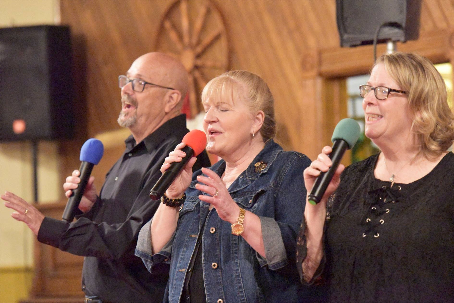 Three people singing into microphones on a stage with a wooden backdrop.