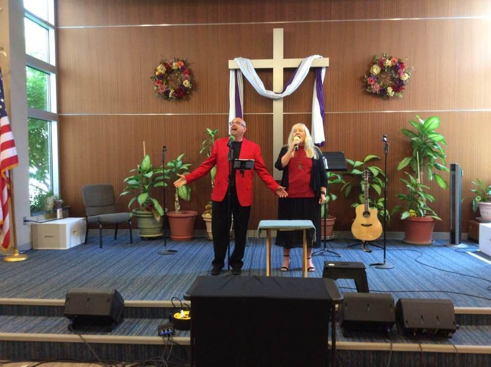 Two singers on stage with a cross, potted plants, and an American flag.