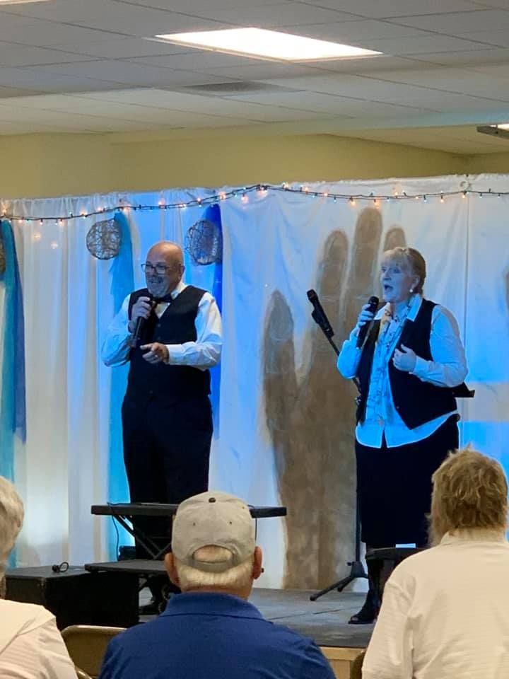 Two people singing on a stage, wearing vests, with a backdrop of a large hand and blue accents.