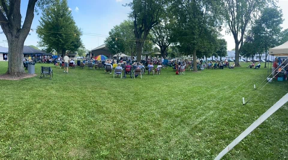 People seated in chairs on grassy park, under trees. Bright, sunny day.
