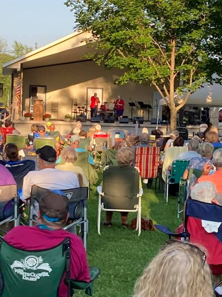 Outdoor concert with audience seated on grass, watching performers on a stage under the sun.