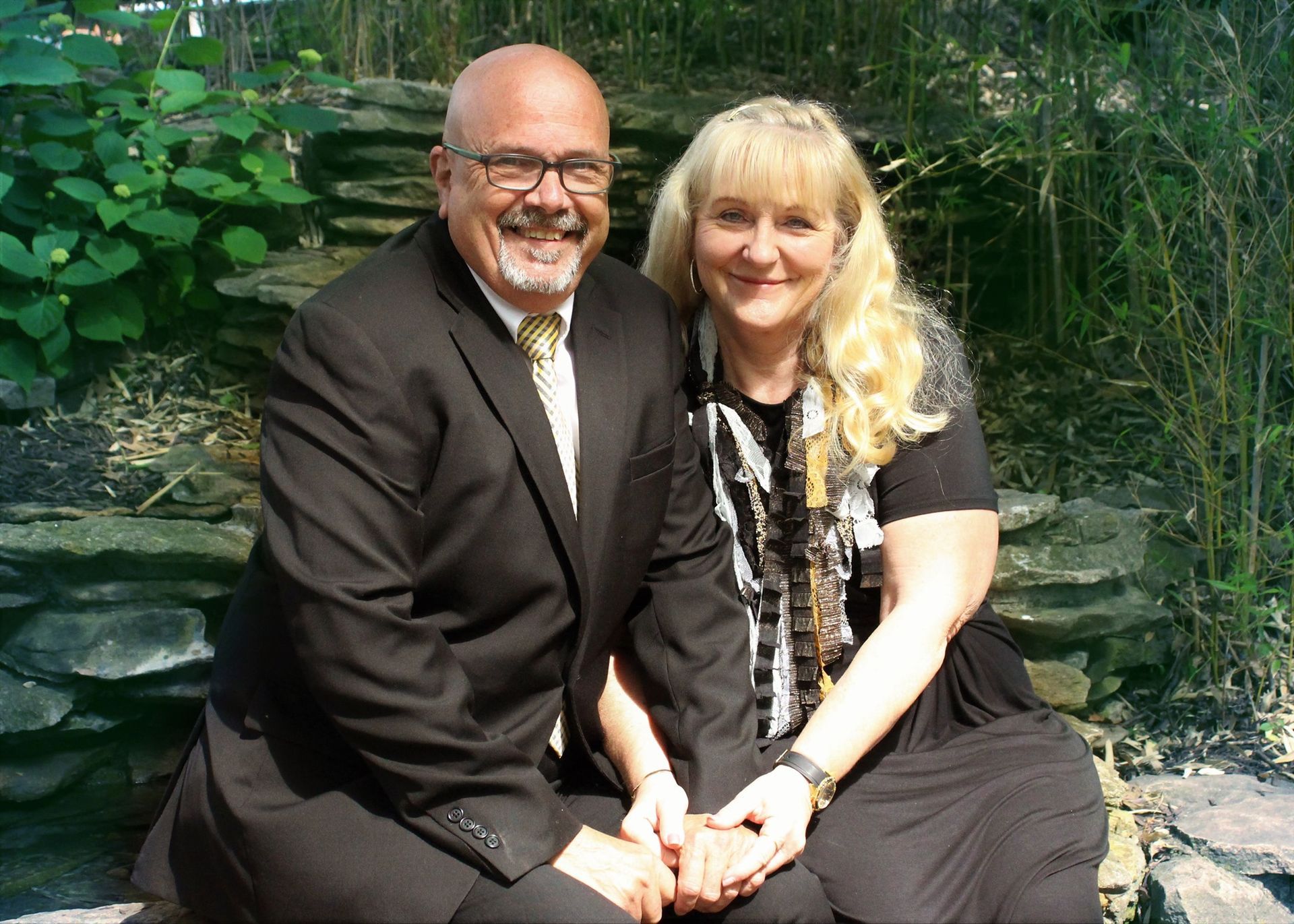 Smiling couple sits close together outdoors; man in black suit, woman in black dress, holding hands.