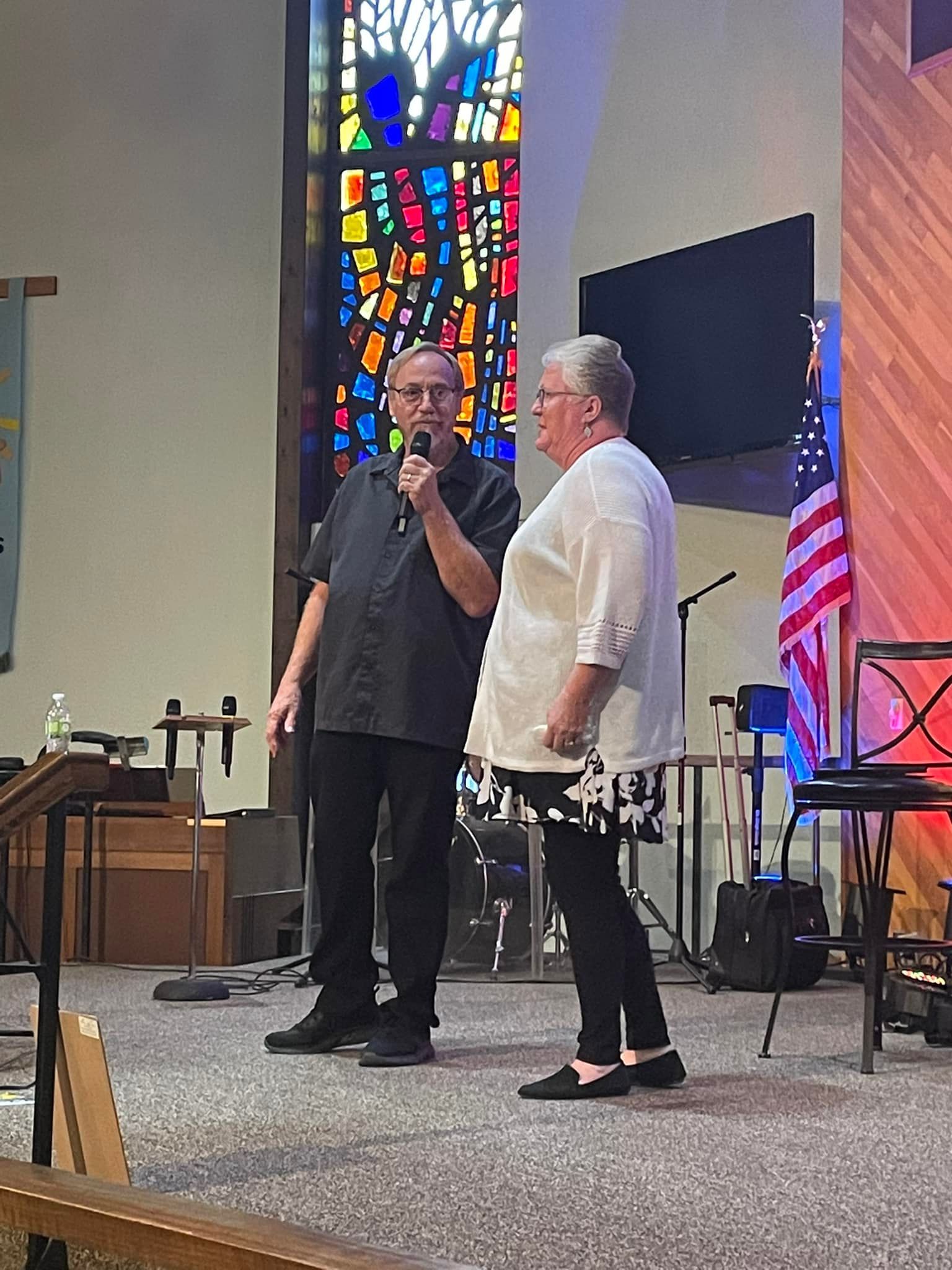 Man speaking into microphone with woman in front of a church stage with stained glass.