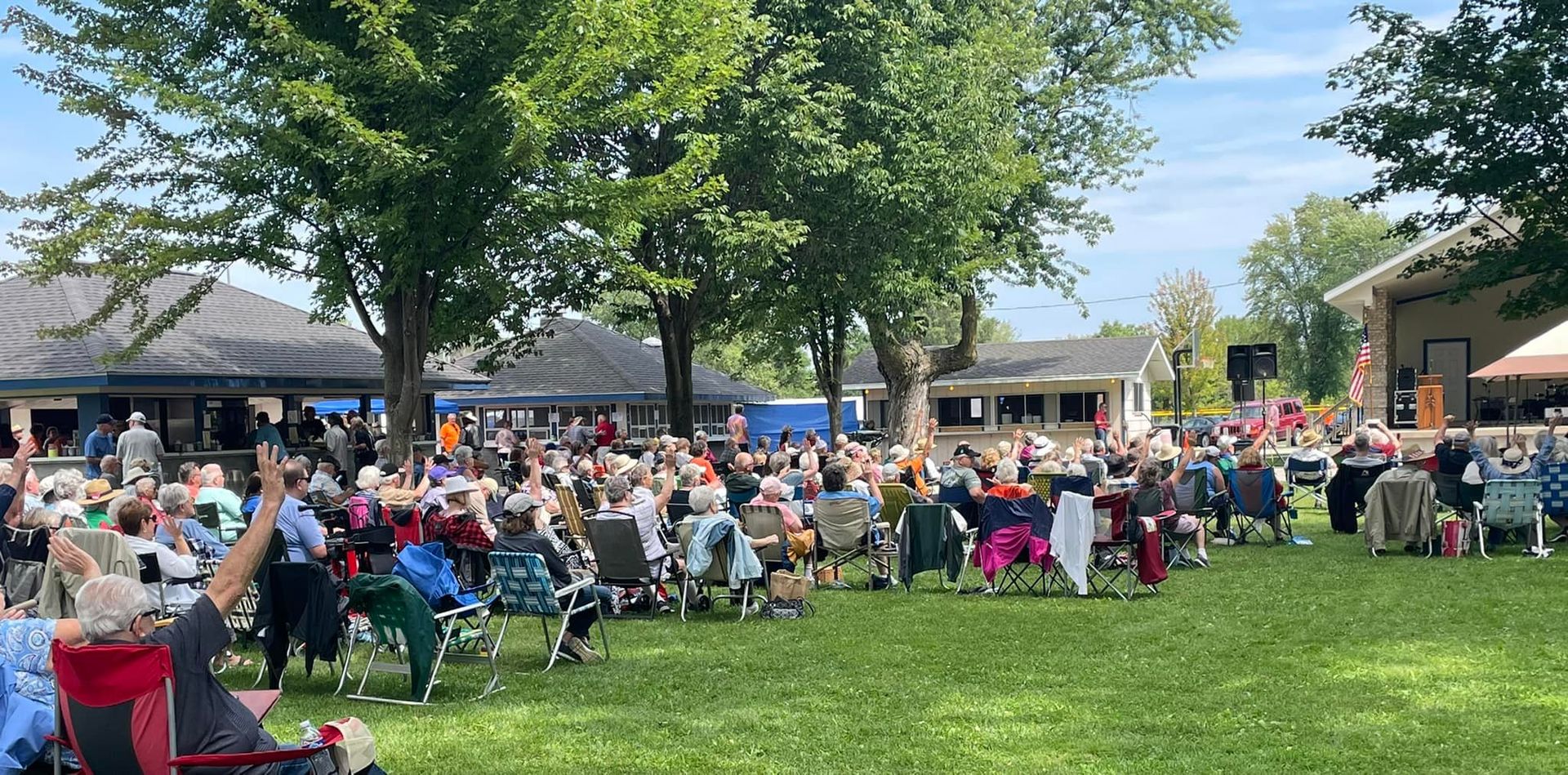 A large crowd sitting in lawn chairs on a grassy area, possibly at an outdoor event on a sunny day.