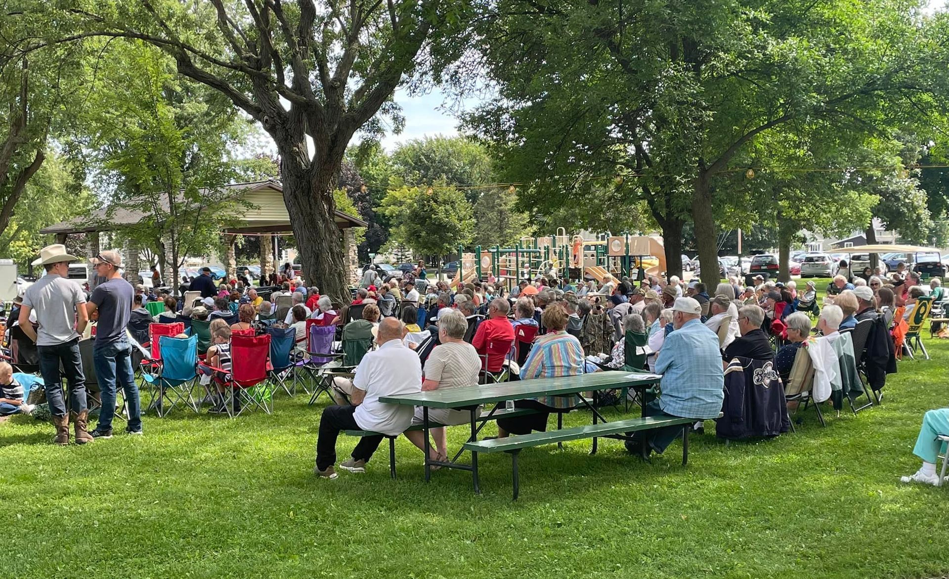 People gathered outdoors in a park. Large crowd seated near picnic tables and folding chairs under trees.