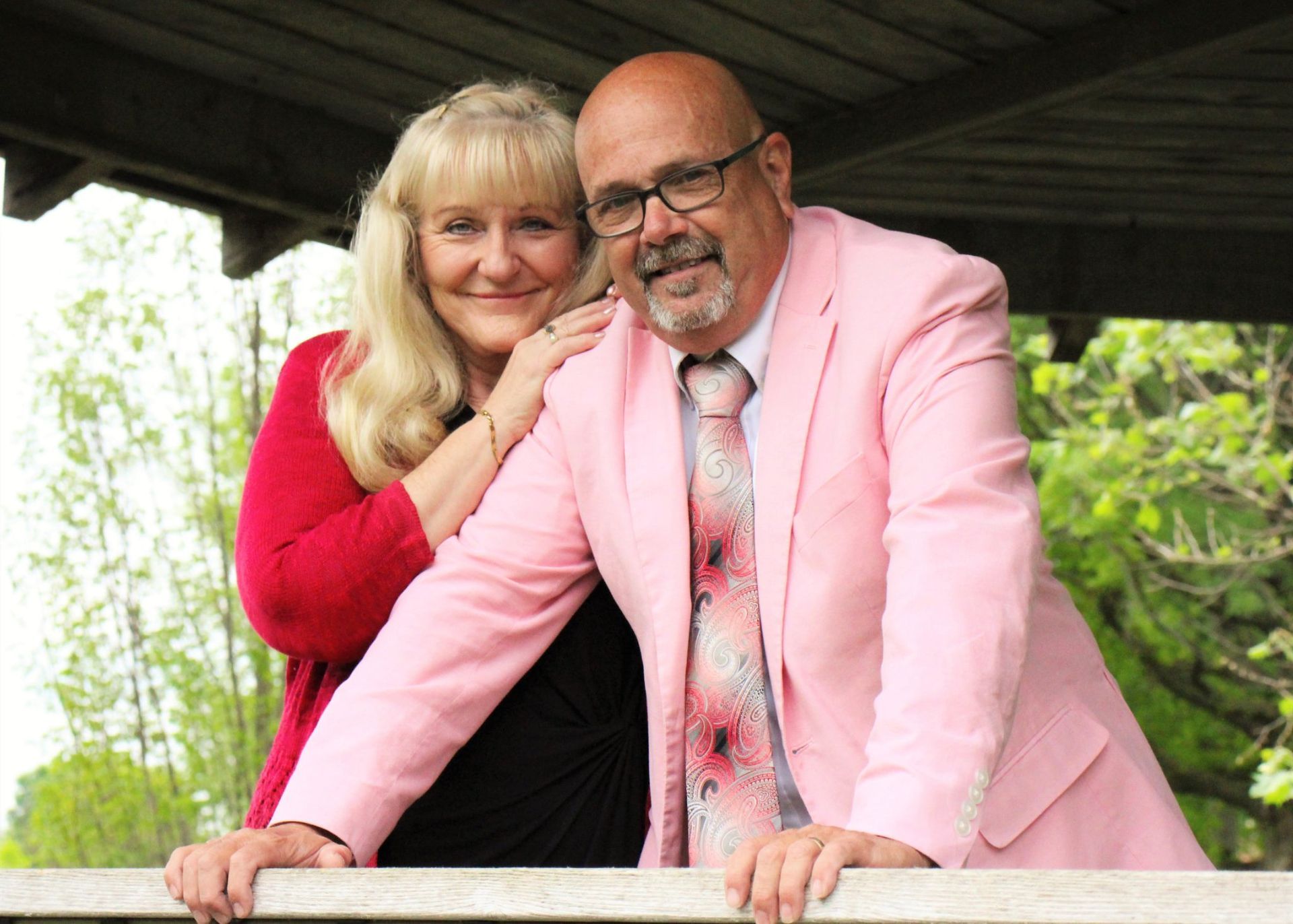 Couple poses smiling outdoors; man in pink suit, woman in red.