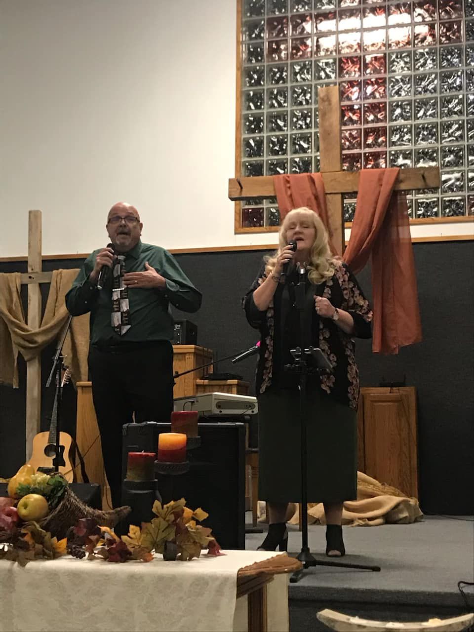 Two people singing into microphones on a stage with a wooden cross and autumn decorations.