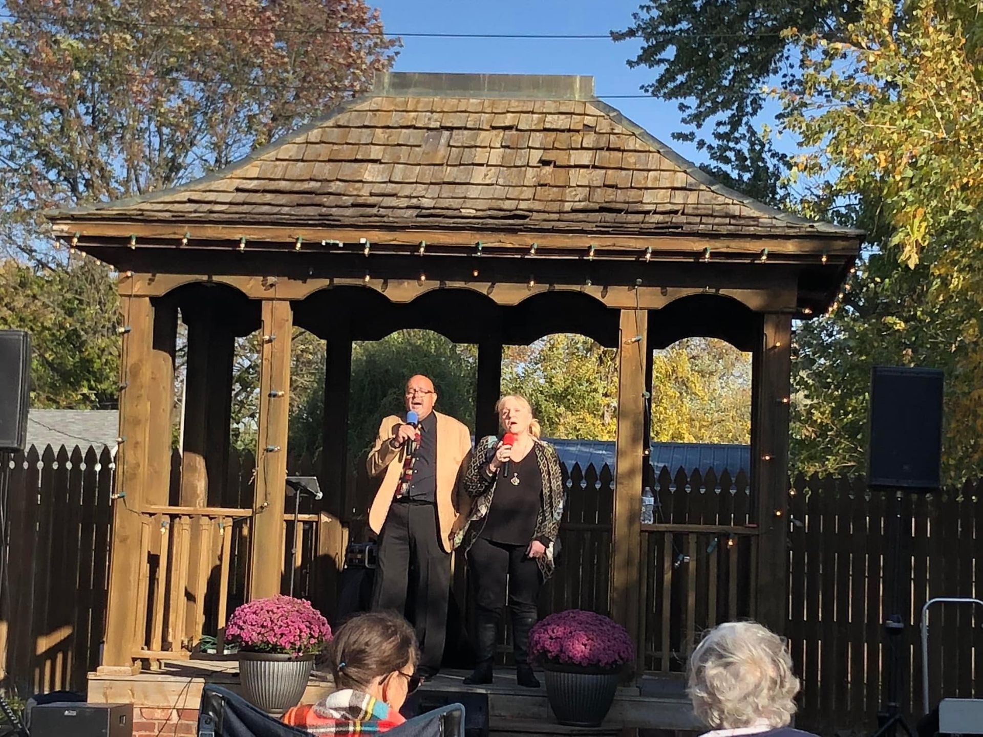Two people sing on a gazebo stage, surrounded by fall colors and an audience.