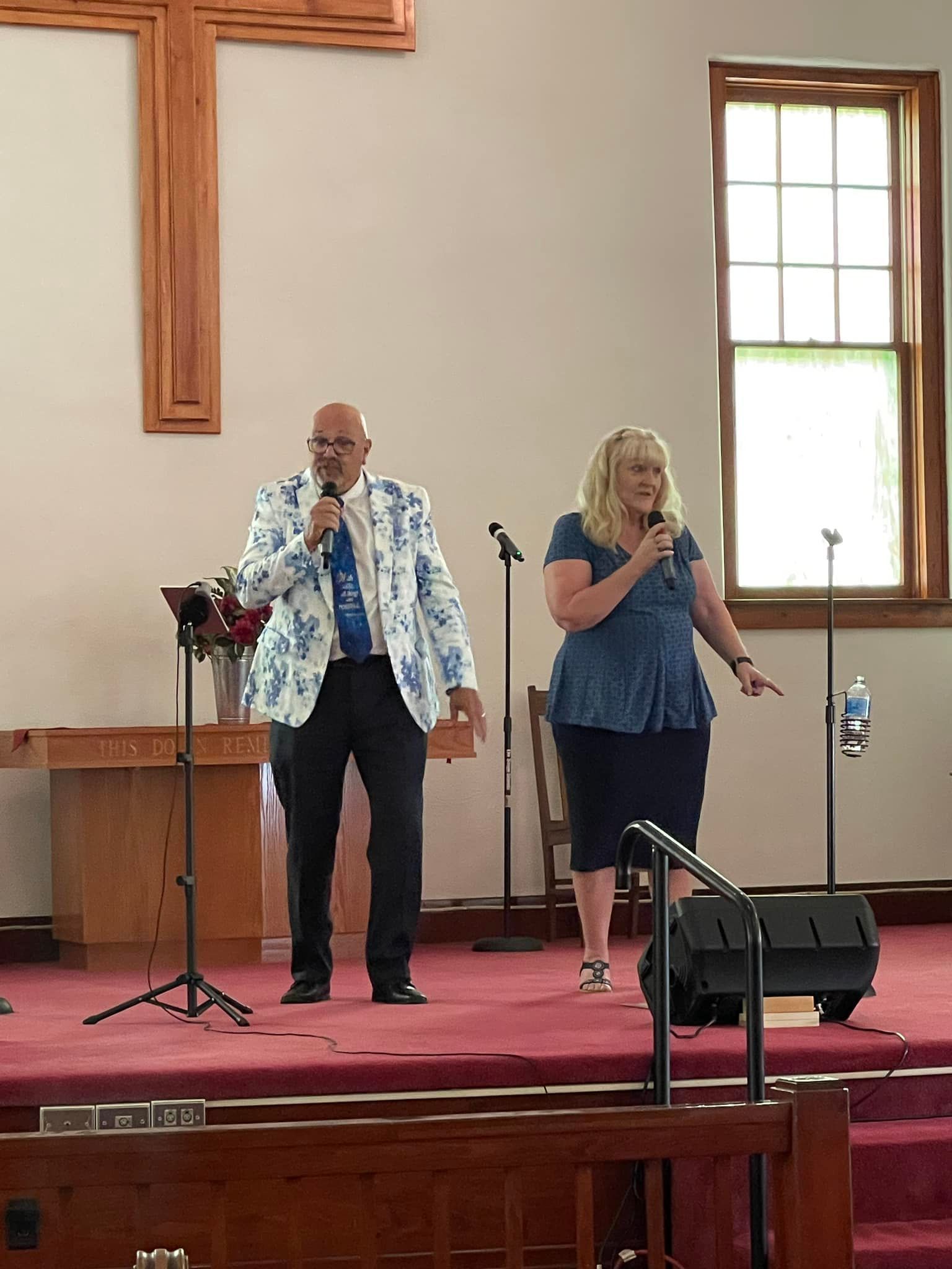 Two people sing into microphones on a church stage; man in blue blazer, woman in blue top.