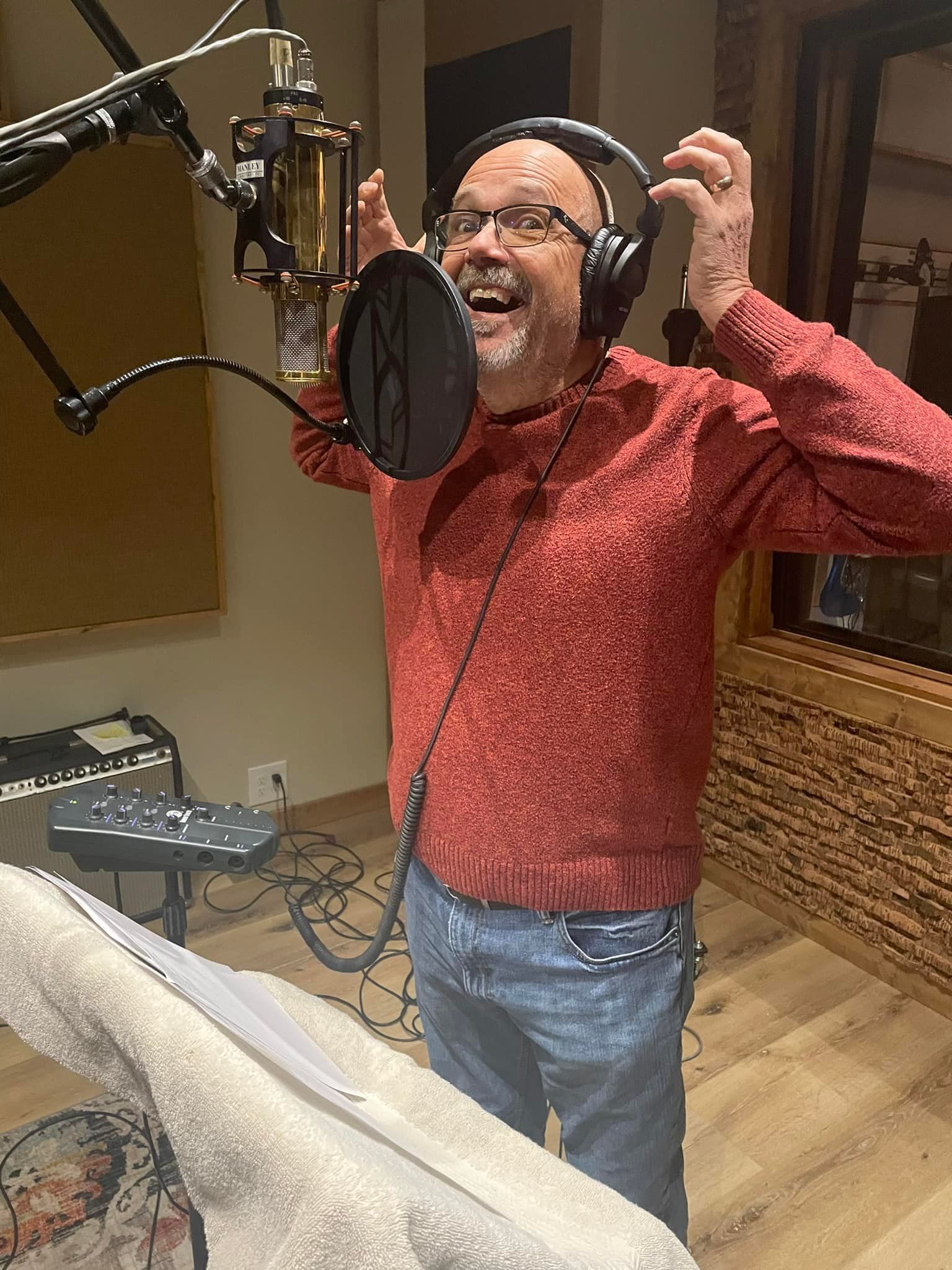 Man in recording studio with headphones, singing into a microphone, red sweater, jeans, joyful expression.