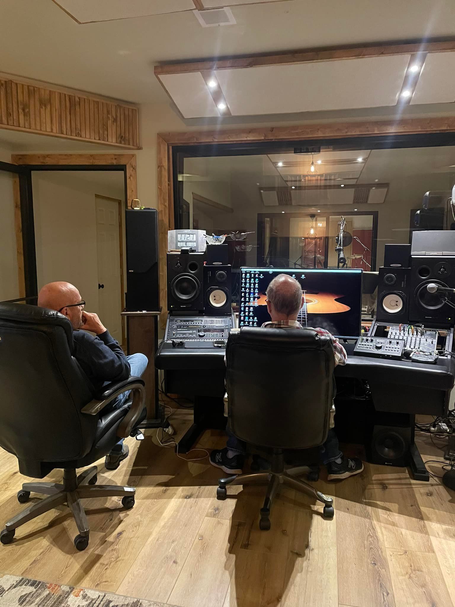 Two people in a recording studio; one sitting, the other in front of the console. Wooden paneling.