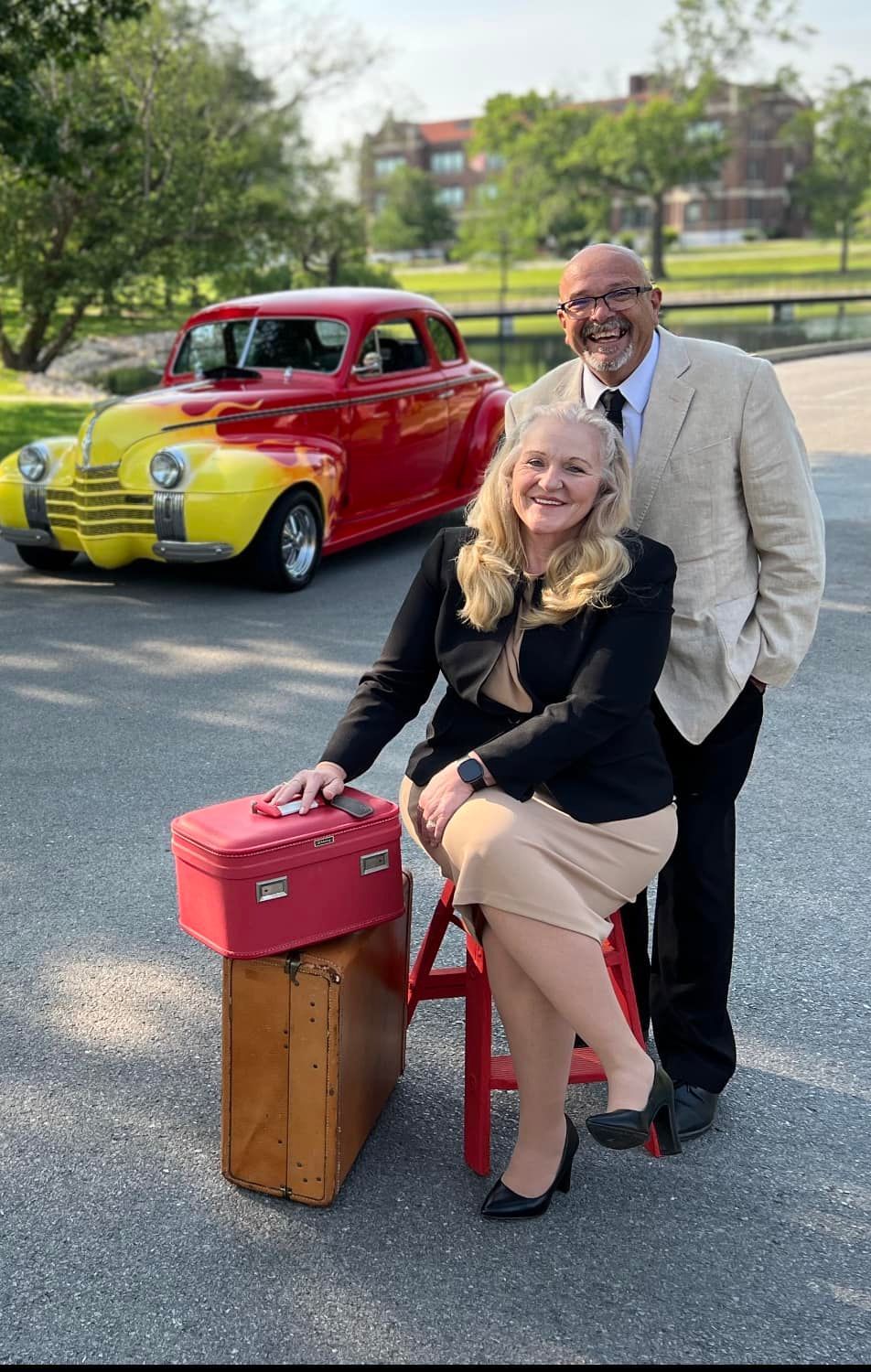 Couple poses by vintage car, woman seated on stool holding suitcase. Man in suit. Red/yellow car.