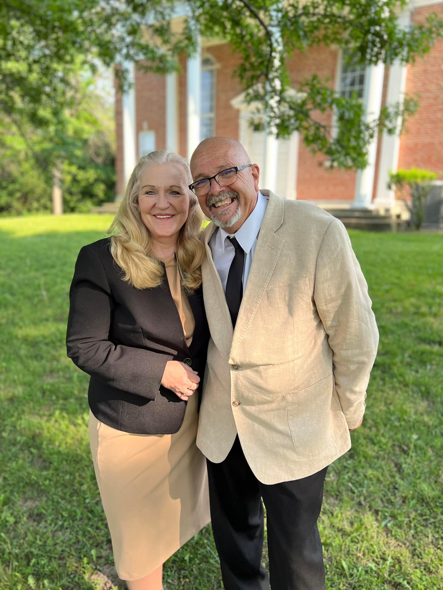 Woman and man smiling, posed outdoors near a brick building. She wears a black blazer, tan skirt. He wears a tan jacket, black pants.