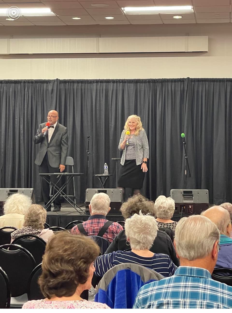 Two people onstage with microphones, entertaining an audience at an event.