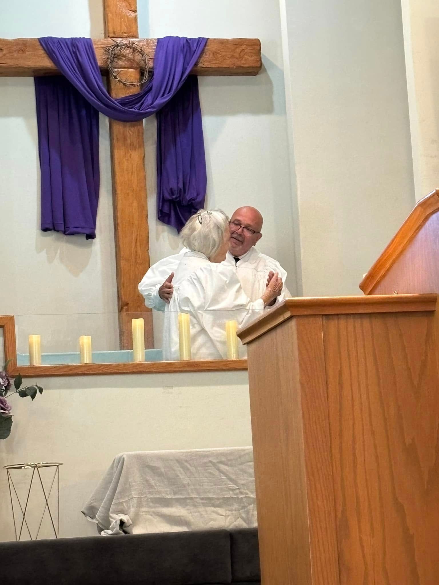 Pastor baptizes a woman in a church, with a cross and purple cloth in the background.