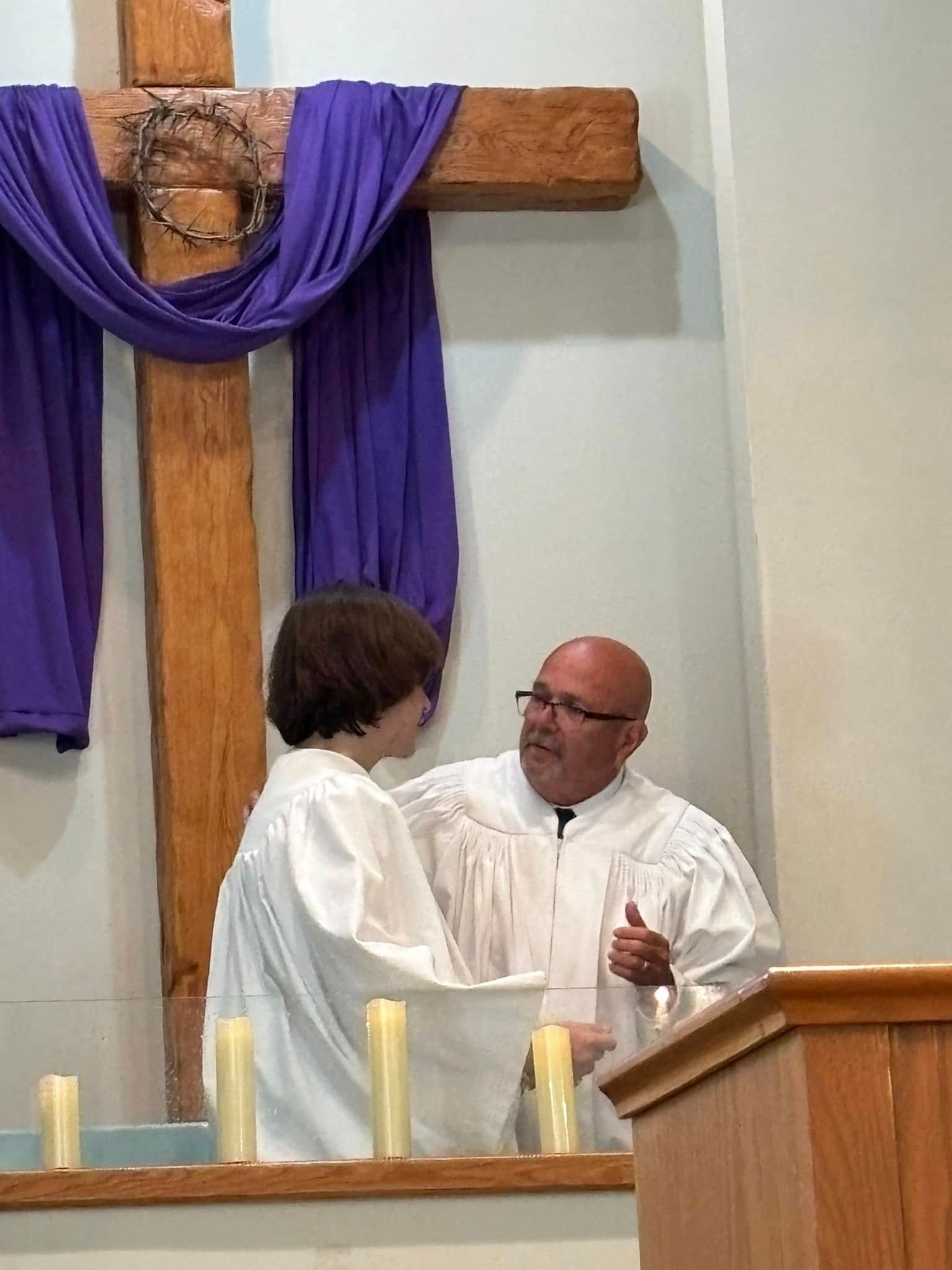 A man in a white robe baptizes a person in white near a wooden cross with purple fabric.