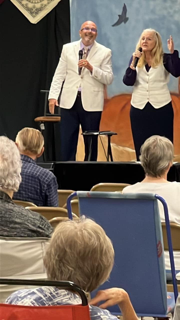 Two people singing on stage, smiling. An audience watches. Stage backdrop features a bird and landscape.