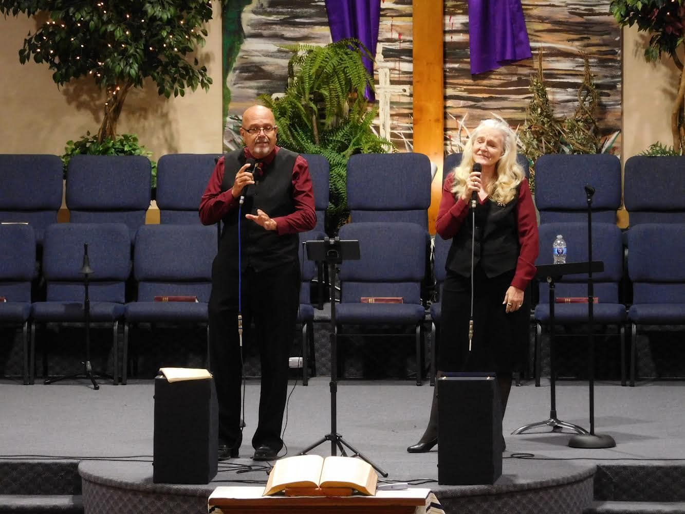 Man and woman singing on a stage in front of blue chairs, both holding mics, with a cross in the background.